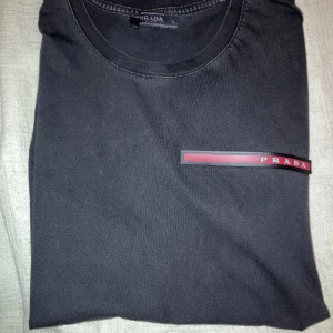 Svart t-shirt från Prada - Svart t-shirt från Prada i bomull med klassisk rund hals och korta ärmar. På bröstet sitter en röd och svart logodetalj med PRADA-tryck. Enkel och stilren design som ger en lyxig känsla.