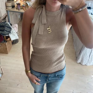 Beige ribbad ärmlös topp med knyt - Supersnygg beige ribbad topp med knyt vid axeln. Toppen är ärmlös och har en tight passform som framhäver figuren. Materialet är stickat med inslag av glittriga trådar som ger en extra touch. Perfekt att styla med jeans eller kjol för en trendig look.