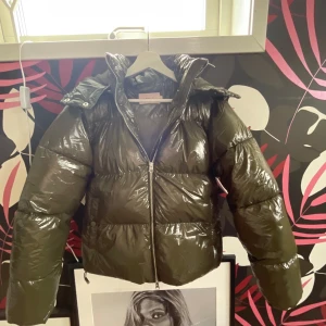 Glansig grön pufferjacka från MbyM - Säljer en riktigt snygg glansig pufferjacka i mörkgrönt från MbyM. Jackan har hög krage, dragkedja framtill och stora puffiga ärmar. Perfekt för kalla dagar när du vill hålla stilen och värmen.