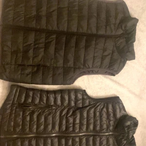 Svart quiltad dunväst med dragkedja - Säljer en svart dunväst med quiltad design och hel dragkedja framtill. Västen har ståkrage och två fickor med dragkedja. Perfekt för lager-på-lager och enkel att matcha med olika outfits.