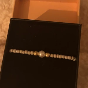 Pärlarmband med vita och guldiga pärlor - Ett stilrent armband med små vita pärlor och två guldiga pärlor som ramar in en större vit pärla i mitten. Perfekt för dig som gillar minimalistiska smycken med en touch av elegans. Elastiskt band gör det enkelt att ta på och av.
