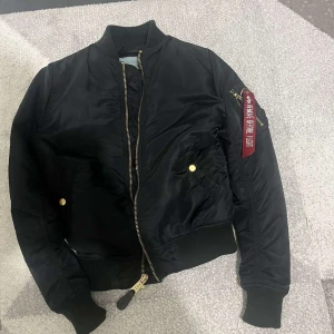 Svart bomberjacka Alpha Industries - Snygg svart bomberjacka från Alpha Industries med klassisk röd tag på ärmen och guldfärgade detaljer. Jackan har ribbade muddar, dragkedja framtill och två fickor med tryckknappar. Perfekt för dig som vill ha en streetig och tidlös look.