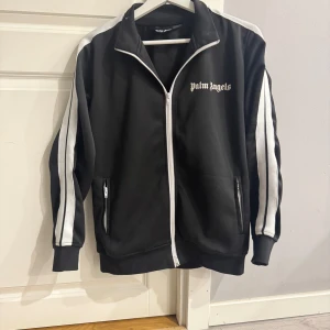 Palm Angels svart track jacket - Svart track jacket från Palm Angels med vita ränder längs ärmarna och vit dragkedja. Klassisk sportig look med hög krage och logga på bröstet. Två fickor med dragkedja och ribbade muddar