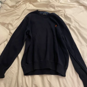 Mörkblå stickad tröja Polo Ralph Lauren - Klassisk mörkblå stickad tröja från Polo Ralph Lauren med rund halsringning och den ikoniska röda logon broderad på bröstet. Tröjan har långa ärmar och ribbade muddar för en snygg passform. Perfekt för dig som gillar stilrena och tidlösa plagg. Passar ungefär S-M