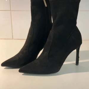 Svarta stövletter med klack - Säljer ett par svarta stövletter i mocka-look med spetsig tå och smal hög klack. Skorna har dragkedja på insidan för enkel påtagning och en elegant, stilren design som passar till många outfits.