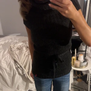 Svart stickad polotröja med knyt - Mysig svart stickad polotröja utan ärmar och med bred krage. Tröjan har ett knytband i midjan som ger en snygg siluett. Perfekt att styla med jeans eller kjol för en chill men ändå uppklädd look.