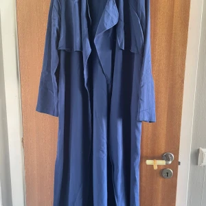 Blå lång trenchcoat med slapp passform - Snygg blå trenchcoat med lång och flowy design. Jackan har bred krage, långa ärmar och ett avslappnat fall. Perfekt för dig som vill ha en stilren och enkel look med lite edge. Passar dig som gillar oversized och trendiga plagg.