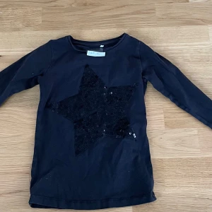 Svart långärmad tröja med paljettstjärna - Svart långärmad tröja med en stor stjärna framtill täckt av svarta paljetter. Tröjan har rund halsringning och är i mjukt bomullsmaterial. Perfekt för dig som gillar lite extra bling i vardagen.