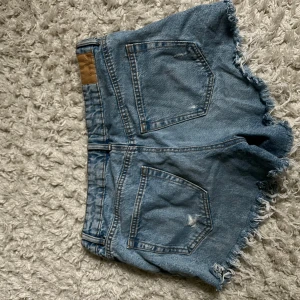 Blå slitna jeansshorts med fransar - Säljer ett par blå jeansshorts från Bershka med råa, fransiga kanter och slitna detaljer framtill. Klassisk femficksmodell med knapp och dragkedja. Perfekta för en avslappnad och trendig sommarlook.