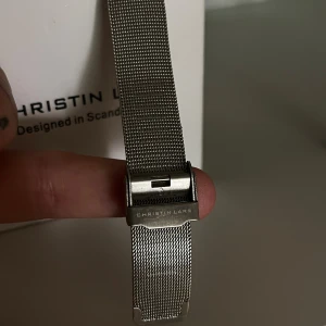 Silverfärgad klocka från Christin Lars - Stilren klocka från Christin Lars med rund urtavla i vitt och silver. Klockan har ett mesharmband i metall och minimalistisk design utan siffror. Quartz-urverk och klassiskt spänne. FYI: batteri behöver bytas.                                                              (Läs alltid min profil för villkor)