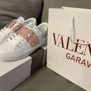 Valentino Rockstud sneakers vit/rosa - Snygga Valentino Garavani Rockstud sneakers i vitt skinn med ljusrosa band och ikoniska nitar. Klassisk rund tå, platt sula och vita snören. Märkeslogga på plös och sula. Perfekta för dig som vill sticka ut med exklusiv streetstyle.