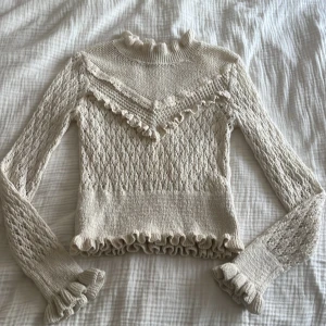 Beige stickad volangtröja från Zara - Supersöt beige stickad tröja från Zara med volangdetaljer vid hals, ärmslut och nederkant. Tröjan har ett genombrutet mönster, lång ärm och en feminin, figurnära passform. Perfekt för dig som gillar romantiska och trendiga vibes.