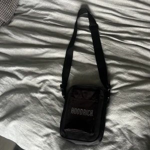Svart axelväska från Hoodrich - Säljer en svart axelväska från Hoodrich med justerbar rem och flera fack. Väskan har tydliga Hoodrich-loggor både fram och bak samt praktisk dragkedja. Tillverkad i slitstark polyester och perfekt för att bära dina viktigaste grejer.
