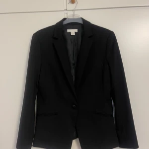Svart kavaj från H&M, storlek 42 - Klassisk svart kavaj från H&M i storlek 42. Enkelknäppt modell med smal krage och två fickor framtill. Snygg passform och helfodrad insida, perfekt till en stilren look. Materialet är mjukt och följsamt, vilket gör den bekväm att bära hela dagen.