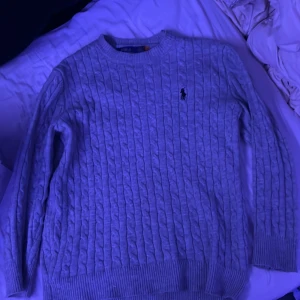 Grå kabelstickad tröja Polo Ralph Lauren - Kabelstickad grå tröja från Polo Ralph Lauren med klassisk rund halsringning och broderad logga på bröstet. Tröjan har ribbade muddar vid ärmslut och nederkant, perfekt för lager-på-lager under kyliga dagar.