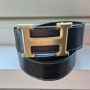 H bälte guld 115 cm - Snyggt svart bälte med ett stort H-format spänne i guld. Bältet är gjort i präglat skinn med vita kontrastsömmar längs kanterna. 115 cm långt, hål-metall stång medföljer d om man vill göra fler hål för att bältet ska sitta bättre ✌️ Hör av dig vid minsta fråga! 🍾 Paketpris vid köp av fler än 1!
