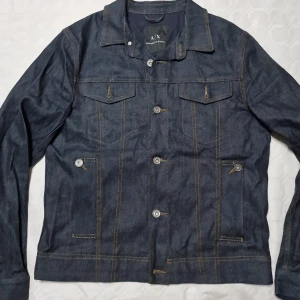 Mörkblå jeansjacka Armani Exchange - Snygg mörkblå jeansjacka från Armani Exchange med klassisk design, kontrastsömmar och knappar framtill. Jackan har två bröstfickor med lock och två sidofickor med knapp. Perfekt för dig som vill ha en stilren och tidlös look.