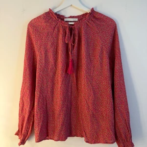 Oui Rosa blus  - Rosa blus med ett mönster i guldbrunt. Blusen har långa ärmar med resår vid handleden och en rynkad halsringning med knytband och en tofs. Storlek 38