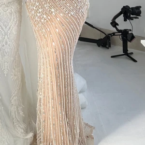 Champagnefärgad glittrig långklänning - Superelegant långklänning i champagnefärg med glittriga paljettdetaljer och pärlbroderier i vertikala mönster. Klänningen har figurnära passform och markerad midja, perfekt för bal eller fest. Materialet känns lyxigt och skimrande.