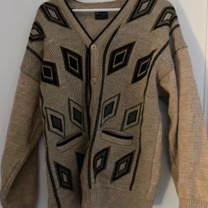 Beige kofta med grafiskt mönster Raja - Beige stickad kofta från Raja med coola grafiska diamantmönster i grönt, svart och brunt. Koftan har v-ringning, knäppning framtill med träfärgade knappar och två fickor. Perfekt för dig som gillar retrovibbar och unika detaljer.