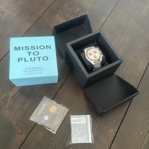 Omega x Swatch Mission to Pluto klocka - Säljer en Omega x Swatch Speedmaster Mission to Pluto från Bioceramic MoonSwatch Collection. Klockan har en rund vit boett, vinröd bezel och vinröda subdials på en beige urtavla. Grått textilband med Omega-logga. Levereras med originalbox och tillbehör.