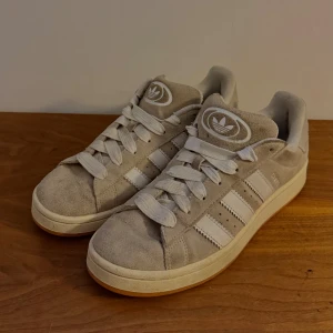 Campus mocka - Säljer ett par beige Adidas Campus sneakers i mocka med klassiska vita ränder och logga på tungan och hälen. Skorna har vit sula och breda vita skosnören. Perfekta för dig som gillar en clean och stilren look med retrovibbar. Är dock en grå fläck på ena hälen.