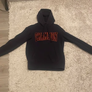 Champion hoodie - Mörkblå hoodie från Champion med stort orange CHAMPION-tryck på bröstet och broderad logga på ärmen. Klassisk huva med snörning och känguruficka framtill. Perfekt för dig som gillar streetwear och vill sticka ut med en fet logga.