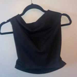 Svart ärmlös topp från Gina Tricot - Säljer en svart ärmlös topp från Gina Tricot med snygg draperad halsringning. Toppen har en croppad passform och är tillverkad i ett mjukt, stretchigt material som sitter bekvämt. Perfekt att styla med jeans eller kjol för en trendig look.