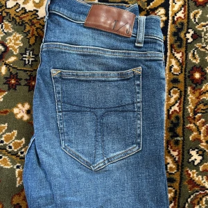 Tiger of Sweden jeans - Säljer ett par blå jeans från Tiger of Sweden med klassisk femficksdesign och snygga slitningar framtill. Jeansen har en slim passform och är tillverkade i mjukt denimtyg med skinnpatch i midjan. Perfekta för dig som gillar en stilren och modern look.
