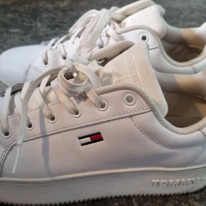 Vita sneakers Tommy Hilfiger - Snygga vita sneakers från Tommy Hilfiger med klassisk logga på sidan och präglad TOMMY-text på sulan. Skorna har snörning, rund tå och platt sula. Tillverkade i skinn och textil för en fräsch och stilren look. Små märken som man nog kan putsa bort. Se bilder.