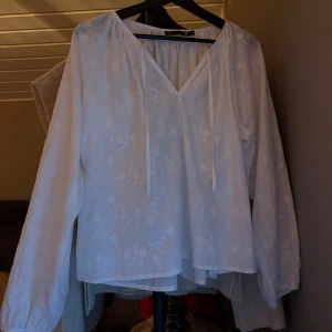 Vit blus med broderade blommor 157 - Luftig vit blus från 157 med broderade blommor och v-ringning. Blusen har långa ärmar, knytband vid halsen och är tillverkad i ett tunt, lätt material som ger en romantisk känsla. Perfekt för vår, höst, vinter och sommar när du vill ha en söt och avslappnad look.🤍