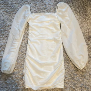 Vit kort klänning från Nelly - Supersnygg vit kort klänning från Nelly med puffiga långa ärmar och fyrkantig urringning. Klänningen har en tight passform med rynkade detaljer längs hela kroppen där bak och är tillverkad i ett glansigt material som ger en lyxig känsla. Använd 1 gång. 150 kr+frakt.