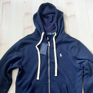 Marinblå zipup Polo Ralph Lauren - Snygg marinblå hoodie från Polo Ralph Lauren med vit broderad logga på bröstet. Tröjan har dragkedja, huva med vita snören och två fickor framtill. Tillverkad i mjuk bomull och fleece på insidan för extra komfort.
