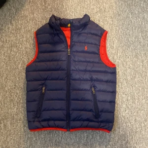 Mörkblå dunväst från Polo Ralph Lauren - Snygg mörkblå dunväst från Polo Ralph Lauren med röda detaljer och röd insida. Västen har två fickor med dragkedja, hög krage och klassisk Polo-logga broderad på bröstet. Perfekt för lager-på-lager och chill stil.