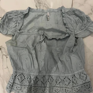 Ljusblå broderad blus från FASSIER - Supersöt ljusblå blus från FASSIER med broderade blommor och volangkant nertill. Blusen har korta puffärmar, knytband i midjan och romantiska detaljer. Perfekt för vår och sommar när du vill ha en luftig och feminin look.