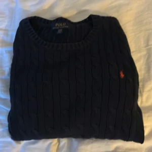Mörkblå kabelstickad tröja Polo Ralph Lauren - Kabelstickad mörkblå tröja från Polo Ralph Lauren med klassisk rund halsringning och orange broderad logga på bröstet. Långärmad modell i mjuk bomull, perfekt för lager-på-lager och snygg till jeans eller kjol.