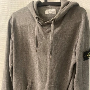 Grå hoodie från Stone Island - Säljer en grå hoodie från Stone Island med klassisk patch på ärmen. Tröjan har huva med snörning, magficka och ribbade muddar. Perfekt för dig som gillar streetwear och vill ha en stilren look.
