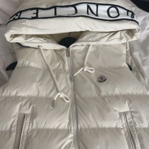 Vit dunväst från Moncler med huva - Snygg vit dunväst från Moncler med stor huva och logga på bröstet. Västen har dragkedja framtill, två fickor med dragkedja och justerbara snören vid huvan. Perfekt för lager-på-lager och streetstyle.