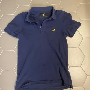 Marinblå pikétröja från Lyle & Scott XS - Marinblå pikétröja från Lyle & Scott i bomull med klassisk krage och korta ärmar. Tröjan har gul broderad logga på bröstet och knäppning framtill. Perfekt för en stilren och sportig look.