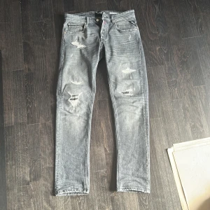 Replay Grover - Säljer ett par Replay Grover jeans i grå tvätt med slitna detaljer och raka ben. Jeansen har klassisk femficksdesign, straight fit och snygga slitningar framtill och på bakfickan. Perfekta för dig som gillar en avslappnad och trendig look.