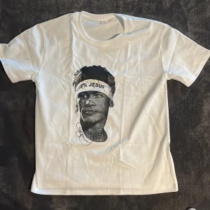Vit t-shirt med grafiskt tryck - Vit t-shirt med korta ärmar och ett stort svartvitt grafiskt tryck av ett ansikte med texten '100% JESUS' på ett pannband. T-shirten har en klassisk passform och är tillverkad i mjuk bomull, perfekt för en avslappnad och cool stil.