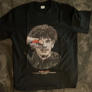 Svart Rockylano t-shirt med tryck - Svart t-shirt från Rockylano i storlek L med ett stort grafiskt tryck på framsidan som visar ett konstnärligt porträtt och textdetaljer. Klassisk rund hals och korta ärmar. Materialet är mjuk bomull som känns skönt mot huden.
