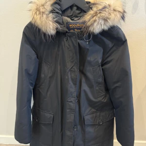 Svart parkas från Woolrich med päls - Svart parkas från Woolrich med stor huva och lyxig pälsdetalj. Jackan har flera fickor framtill, knappar och dragkedja. Perfekt för kalla dagar. Storlek S.