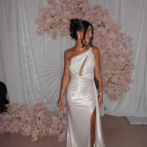 Fest/balklänning - Säljer en elegant champagnefärgad långklänning med pärlemor finish. Klänningen har oneshoulder-design, v-ringning och en hög slits framtill. Materialet är glansigt och ger en lyxig känsla, perfekt för bal eller fest. Insydd till en 34 men går att återgå till 36a om önskat! Priset går att diskutera 