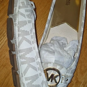 Michael Kors Fulton Moc loafers - Snygga loafers från Michael Kors i modellen Fulton Moc. Skorna är vita med bruna detaljer och har ett diskret MK-mönster över hela ytan. Framtill sitter en guldfärgad MK-logga. Tillverkade i PVC-material och har en platt sula för skön komfort.