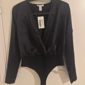 Svart omlott body i satin från NLY One - Snygg svart body från NLY One i satin med omlott-design och djup v-ringning. Långa ärmar och åtsittande passform som ger en stilren och trendig look. Perfekt att styla med höga jeans eller kjol för en cool outfit.
