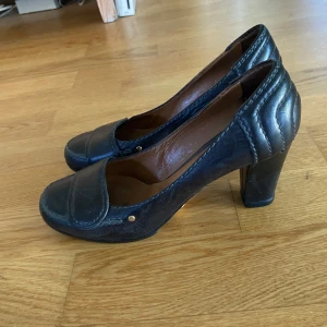 Svarta pumps/ klackskor från Chloé i skinn - Snygga svarta pumps från Chloé i äkta skinn med markerade sömmar och chunky klack. Skorna har rund tå och diskreta gulddetaljer vid sidan. Insidan är fodrad i brunt läder för extra komfort. Perfekta för dig som gillar klassisk men cool vintage stil.