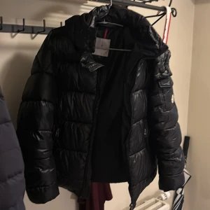 Svart dunjacka från Moncler+NFC tag - Snygg svart dunjacka från Moncler med huva och dragkedja. Jackan har ett stilrent märke på ärmen och praktiska fickor med dragkedjor. Perfekt för kyliga dagar. Använd 1 gång, säljer den för den är för stor. S men passar M+. Kan gö byte med en moncler cardigan 
