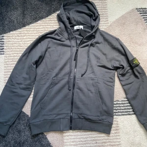Grå hoodie från Stone Island L - Snygg grå hoodie från Stone Island med dragkedja, huva och klassisk logga på ärmen. Tröjan har känguruficka framtill och justerbara snören i huvan. Perfekt för dig som gillar streetwear och vill ha en stilren look.