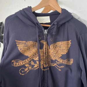 Mörkblå hoodie från Ralph Lauren - Snygg mörkblå hoodie från Ralph Lauren Denim & Supply med dragkedja och huva. Framsidan har ett stort guldigt tryck med örn och text. Tröjan har snörning i huvan och är långärmad, perfekt för en chill och avslappnad stil.